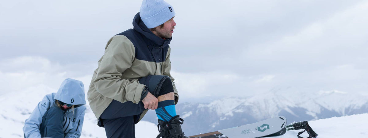 Skier wearing a Bauerfeind knee support fastening a ski boot on a snowy summit: how to prepare your knees for skiing - Bauerfeind UK
